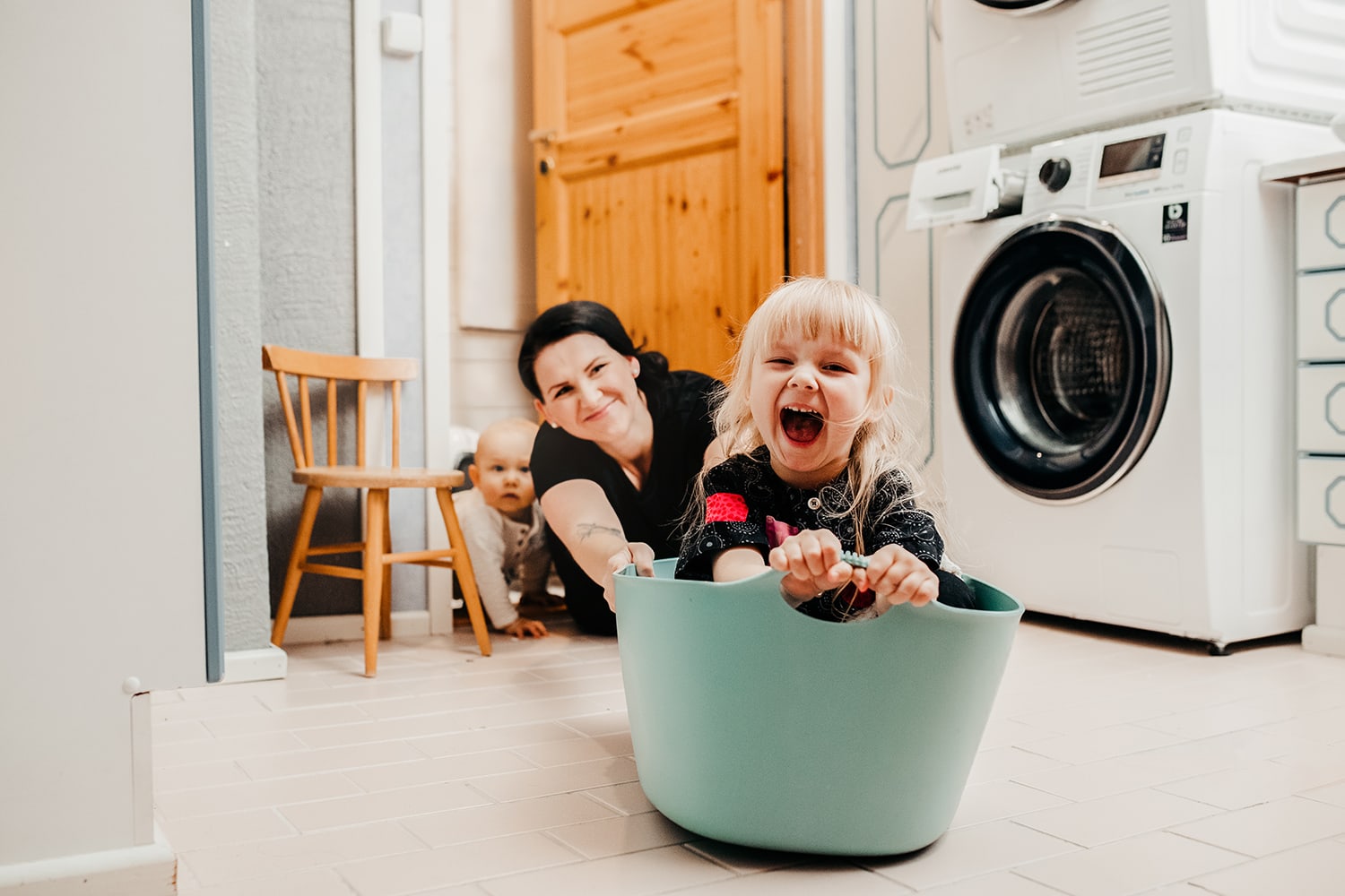 Iloinen lapsiperhe kodinhoitohuoneessa. Pieni naurava tyttö pyykkikorissa.